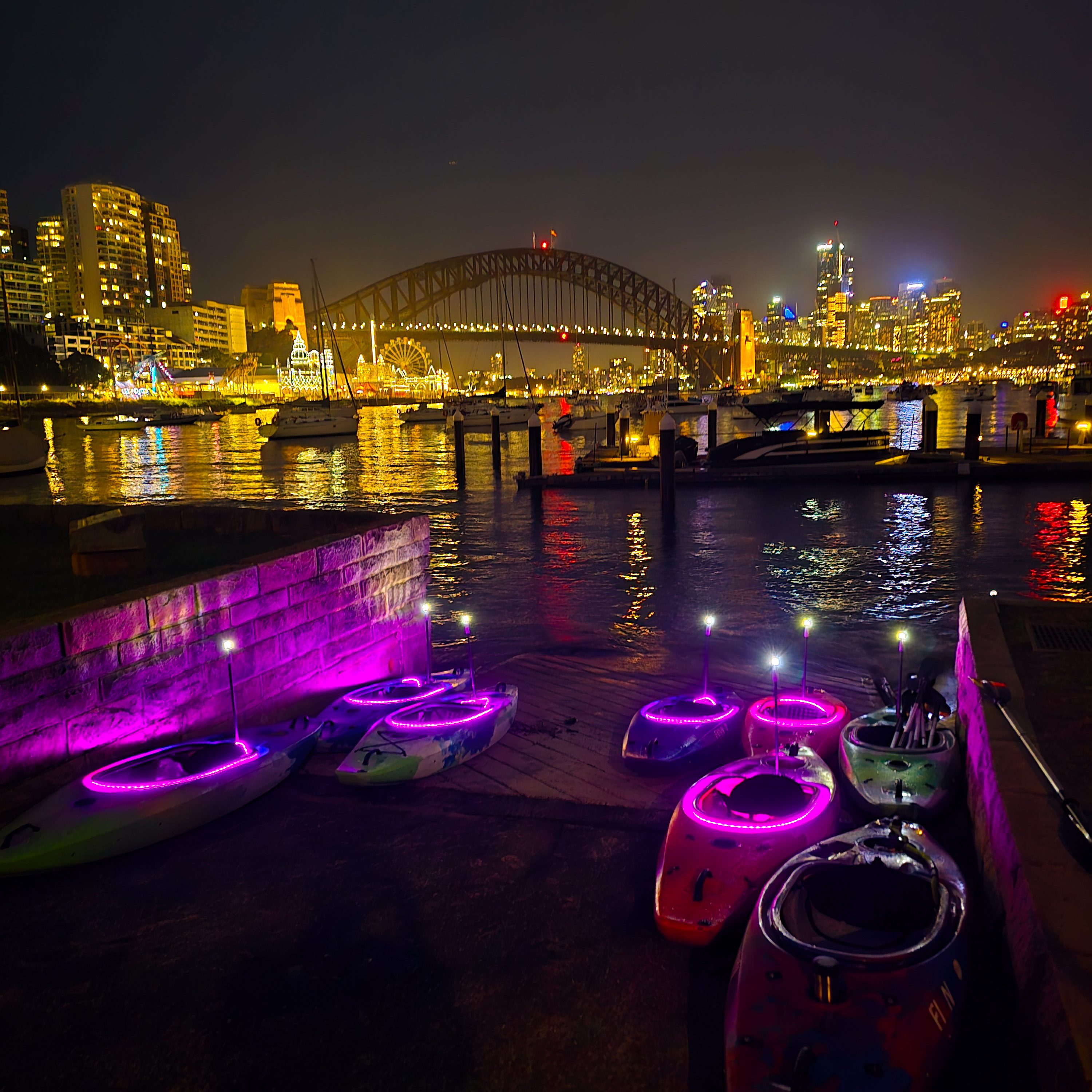 Pink LED-lit kayaks on Sydney Harbour at night with the Opera House and city lights glowing behind