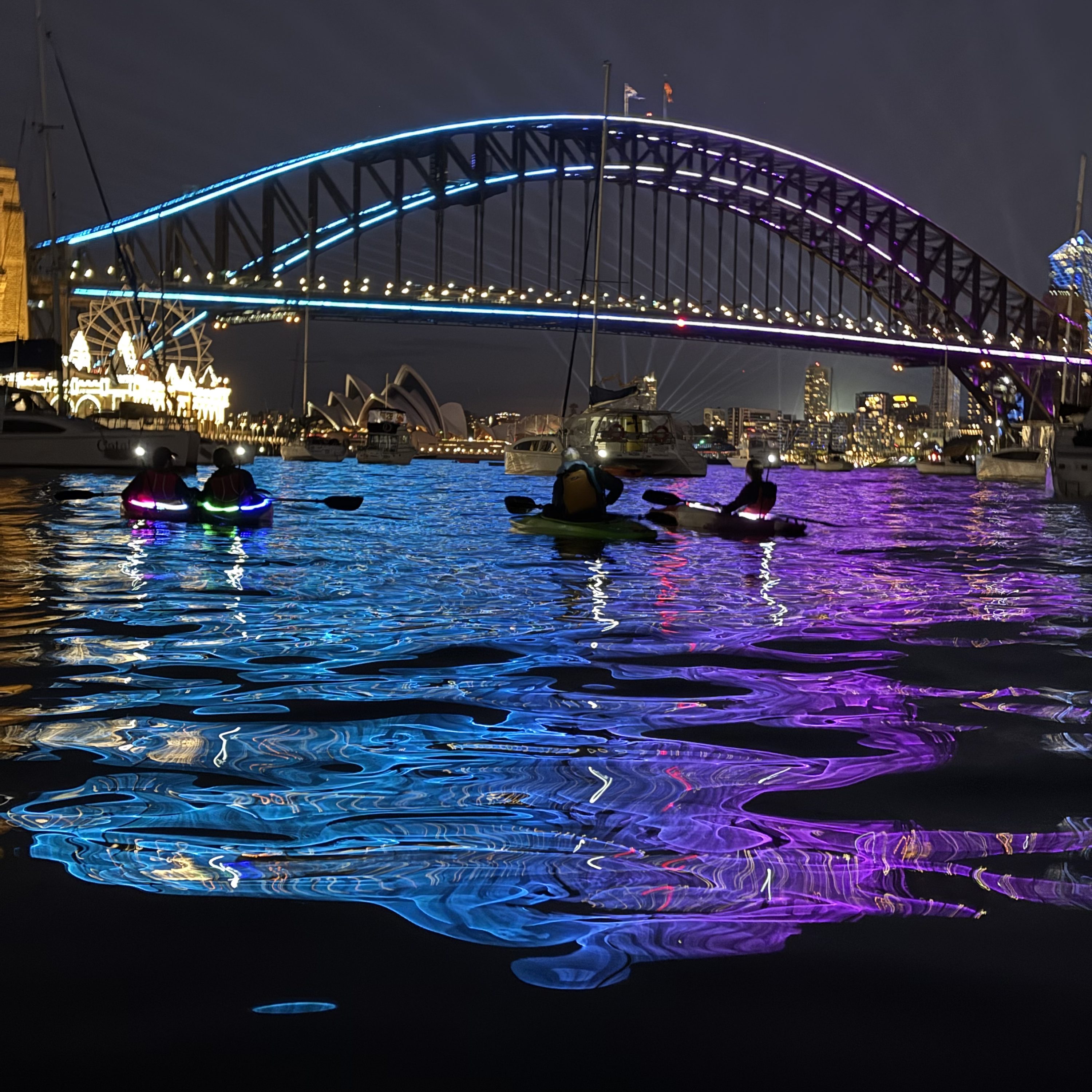 Group of pink LED-lit kayaks on Sydney Harbour at night below the Harbour Bridge
