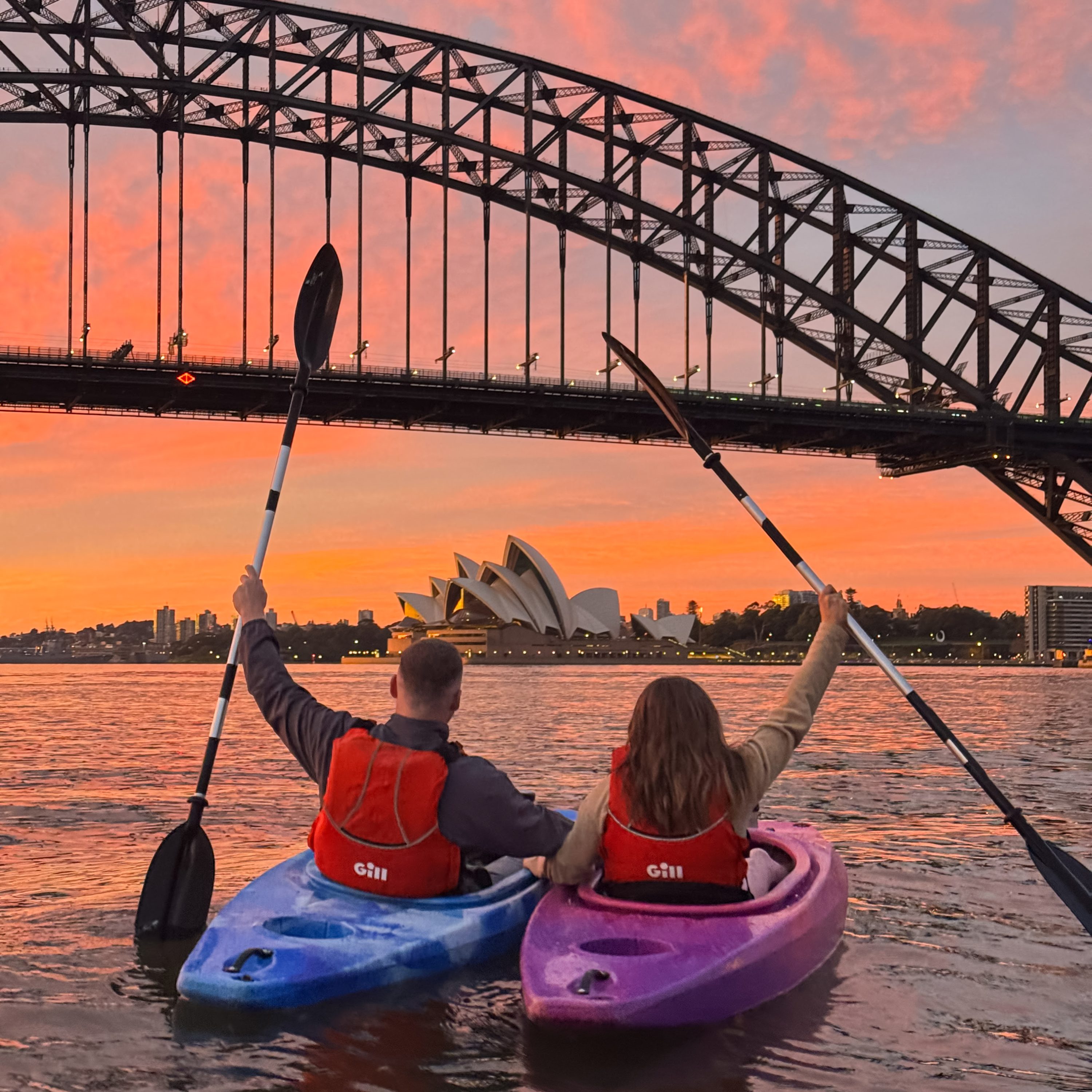 Sydney Harbour Bridge and Opera House silhouetted against a vivid orange sunset, viewed from a kayak on the water