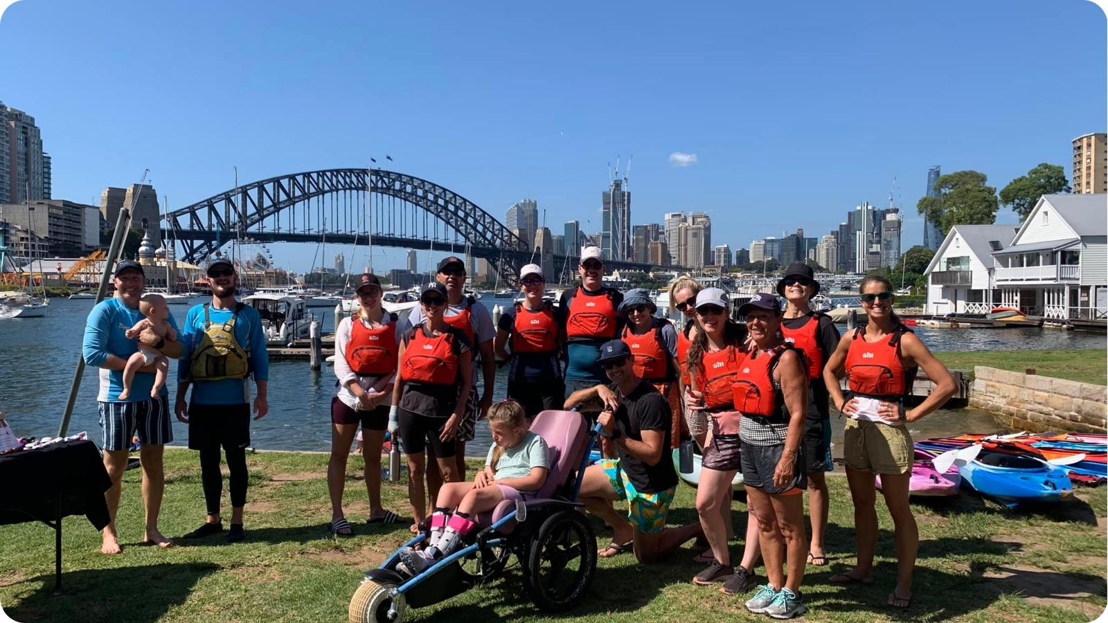 Sydney By Kayak tour group in red life jackets gathered on the grass around girl in wheel chair at Lavender Bay with the Harbour Bridge behind