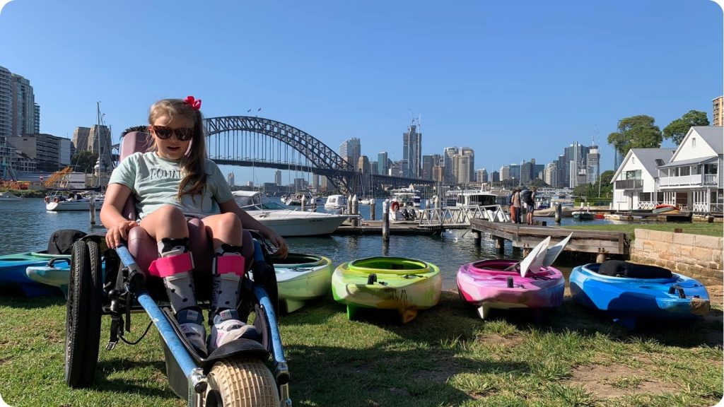 Young girl in a wheelchair beside a row of colourful kayaks at Lavender Bay with the Harbour Bridge behind