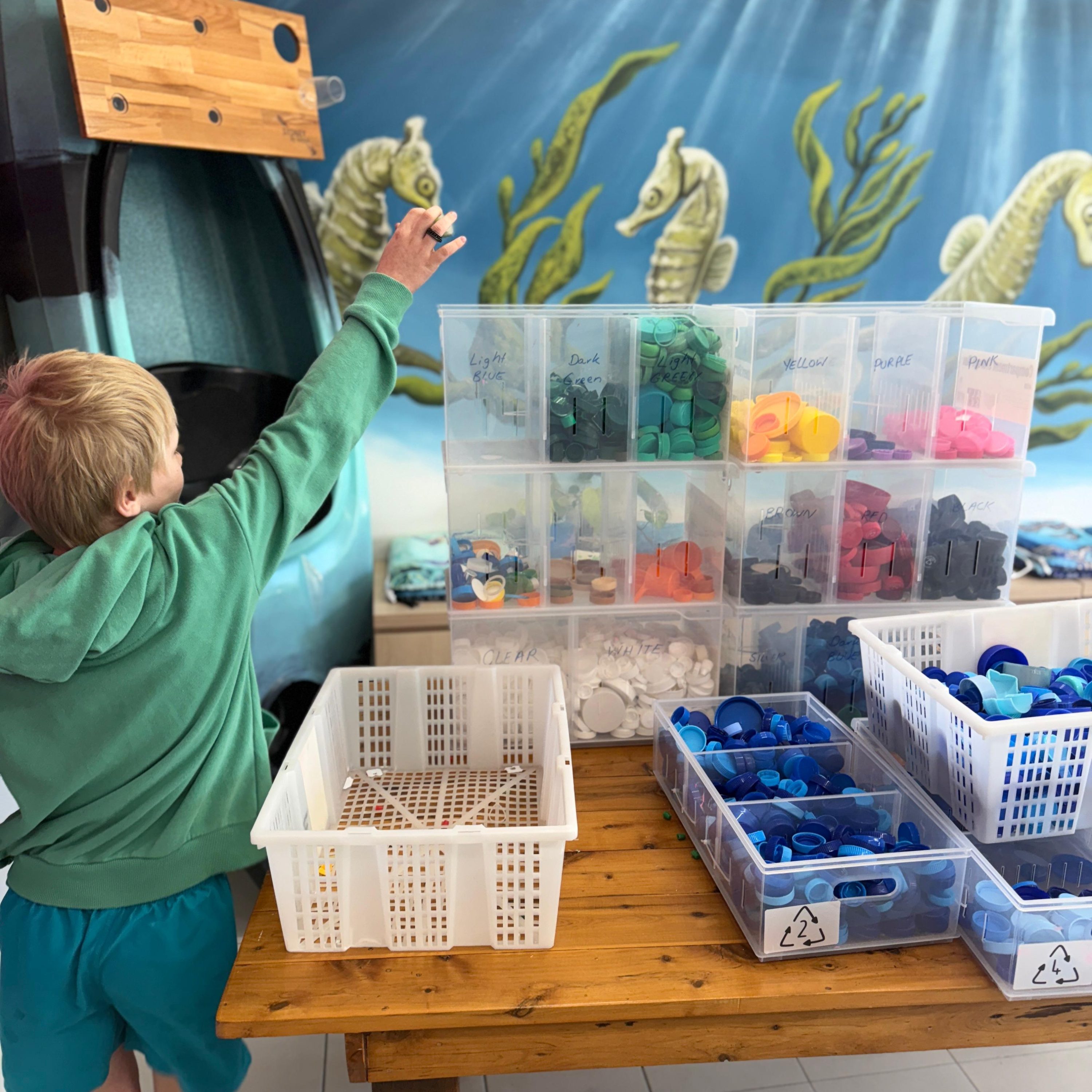 Boy holds up a bottle cap next to a stack of bttle caps inside SBK Eco Hub