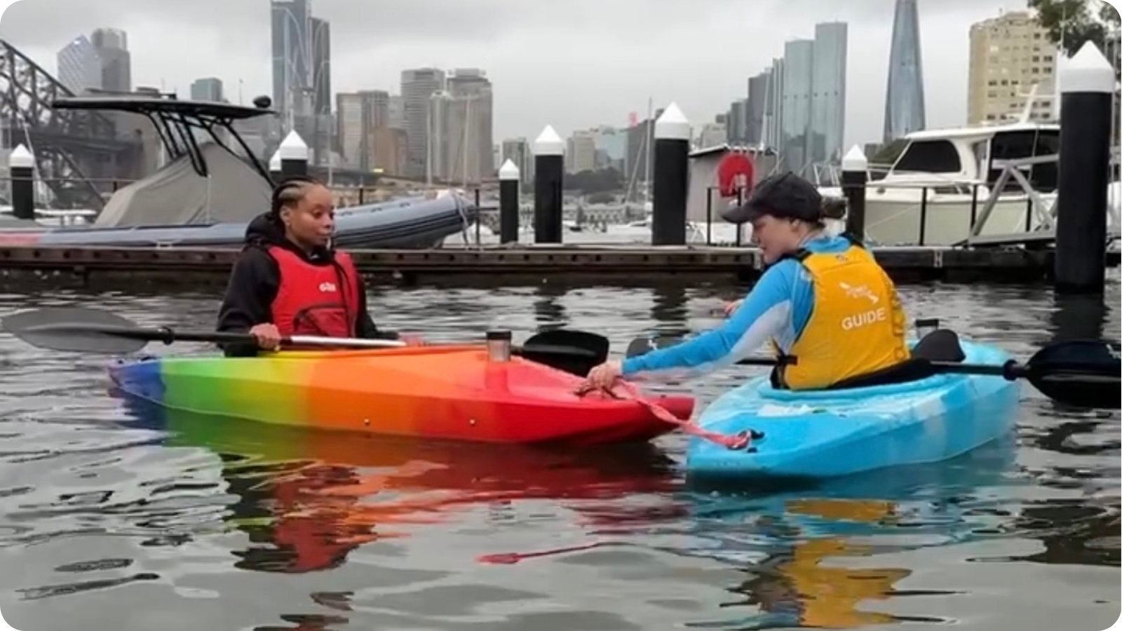 Two paddlers in colourful kayaks on Sydney Harbour with the city skyline behind and one being towed.