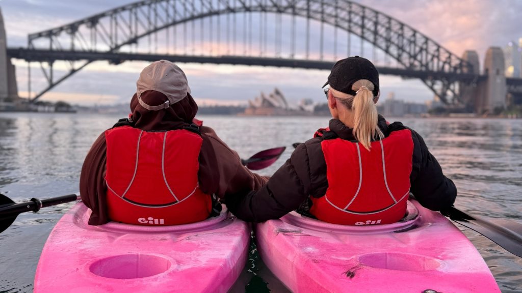 Two paddlers in pink kayaks looking towards the Sydney Harbour Bridge and Opera House at dawn