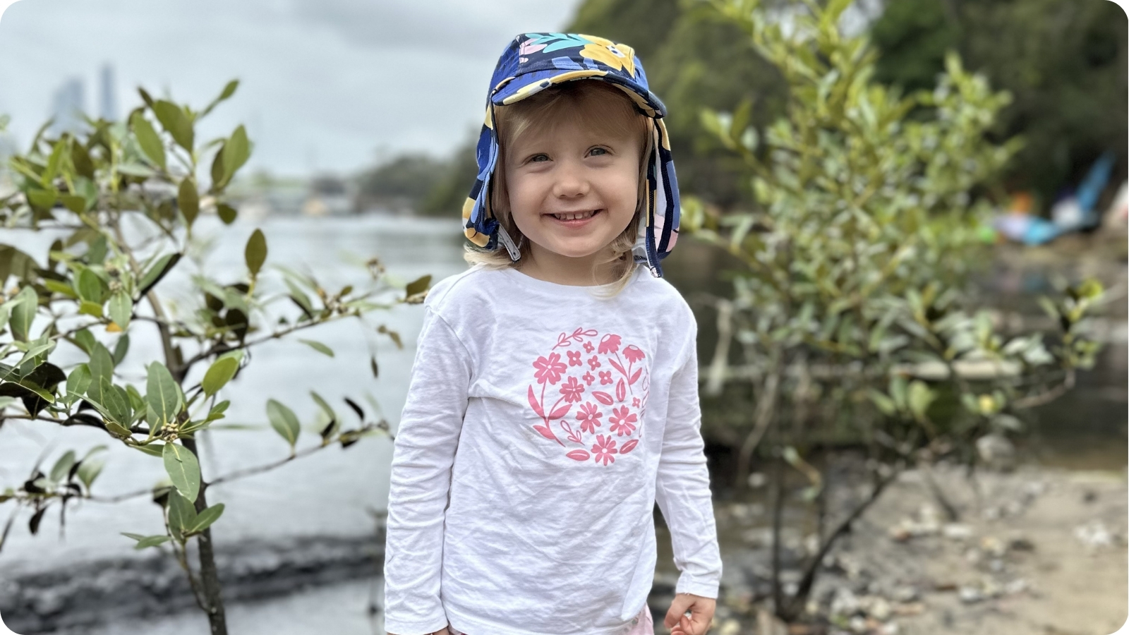 A young child in a sun hat smiling beside mangrove seedlings on the shoreline.