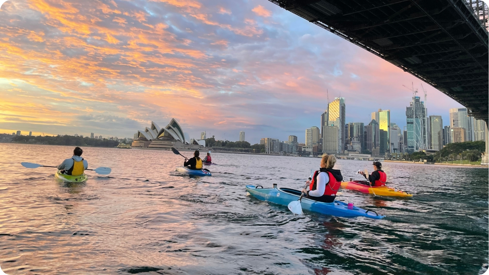 Paddlers heading under the Harbour Bridge at dawn with the city skyline and Opera House ahead.